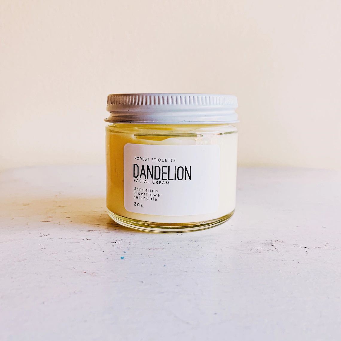 A clear jar with a white lid is filled with a light yellow cream.  The white label on the front reads Dandelion in black lettering